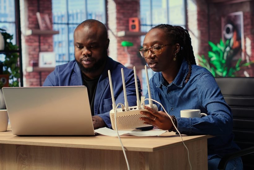 African American couple at home office troubleshooting their Wi Fi modem and discussing solutions to fix a weak connection affecting their remote work productivity. Router guide files. Camera B.